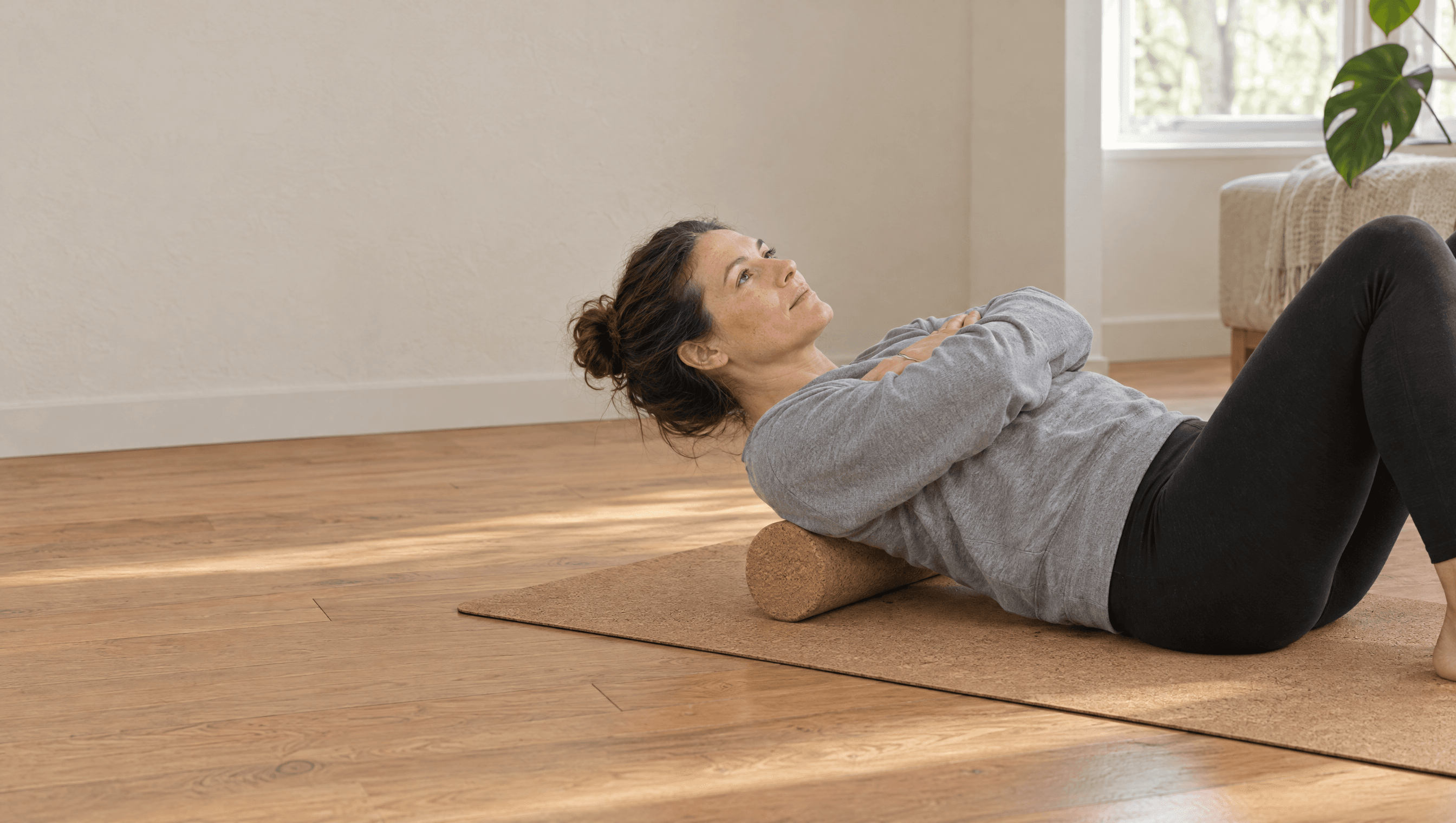 A woman lies on her back on a cork mat with a cork roller under her shoulders in a sunny living room.